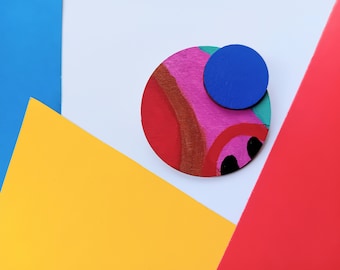 Könnte beinhalten: Eine kreisförmige Holzbrosche mit einem handbemalten Design aus abstrakten rosa, roten und braunen Formen, gekrönt von einem kleineren, einfarbig blauen kreisförmigen Holzstück. Die Brosche weist eine sichtbare Holzmaserung auf der Oberfläche auf.