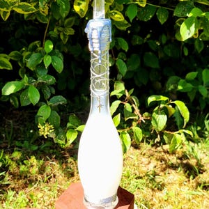 Peut inclure: Une grande bouteille en verre transparent avec un intérieur blanc, surmontée d'une pointe de cristal transparente et de cire bleue. La bouteille est décorée de fil d'argent et d'un petit charme. La bouteille est exposée sur un support en bois marron.