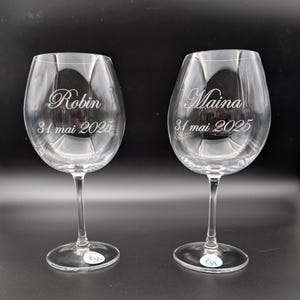Puede incluir: Dos copas de vino de vidrio transparente con los nombres "Robin" y "Maina" grabados en ellas, junto con la fecha "31 mai 2025".