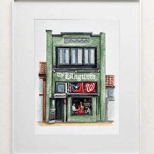 May include: A framed watercolor illustration of a green brick building labeled The Blaguard. The facade features a large window displaying people inside, a dark door, and two red and blue sports flags hanging above the storefront window.