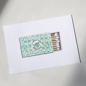May include: A matchbox with a green and white geometric pattern and the text "LA BONNE VACHE GEORGETOWN". The box is open, revealing several wooden matches with white tips. The matches are on a white surface.