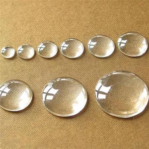 May include: A collection of clear, round glass cabochons of varying sizes. The cabochons are arranged in rows on a tan surface, reflecting light. These glass pieces are suitable for jewelry making or craft projects.