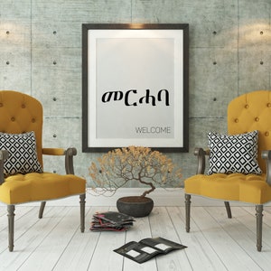 May include: Two yellow chairs with black and white patterned pillows sit in front of a framed print with the text "Welcome" and the Amharic word for "Welcome" in black lettering. A small bonsai tree sits on the floor in front of the chairs.