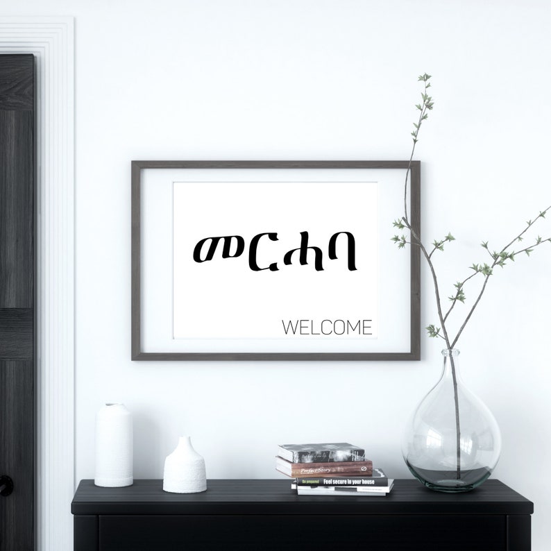 May include: A framed print with the Amharic word for "welcome" in black text on a white background. The frame is brown wood and the print is hanging on a white wall. The word "WELCOME" is written in black text below the Amharic word.