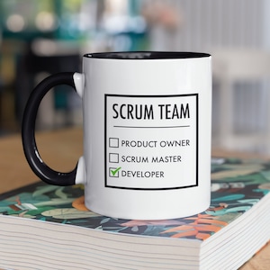 Op de afbeelding: Een witte keramische mok met een zwarte handgreep en een zwarte rand. De mok heeft een zwart-witte afbeelding met de tekst "Scrum Team" met een checklist. De checklist heeft drie items: "Product Owner", "Scrum Master" en "Developer". Het vakje "Developer" is aangevinkt.