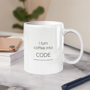 Op de afbeelding: Witte keramische mok met de zwarte tekst "I turn coffee into CODE". De mok heeft een gebogen handvat en staat op een wit marmeren oppervlak. Een smartphone en een stapel wit papier zijn zichtbaar op de achtergrond.