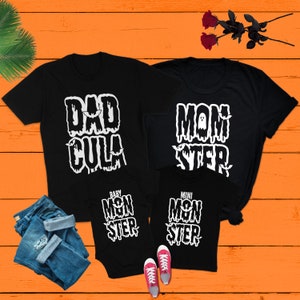 May include: A set of four black t-shirts with a Halloween theme. The shirts feature the text "Dadcula", "Momster", "Baby Monster", and "Mini Monster" in white with a dripping effect. The shirts are on a wooden surface with a pair of pink sneakers and blue jeans.