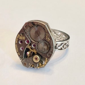 May include: A silver ring with a decorative filigree band and a watch movement as the face. The watch movement has a gold-colored winding stem and red jewels.