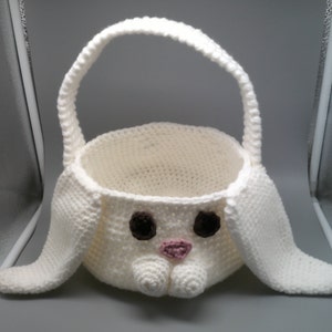 Könnte beinhalten: Ein weißer gehäkelter Osterkorb in Form eines Hasen. Der Korb hat einen runden Körper, lange Ohren und einen Henkel. Das Hasengesicht hat braune Augen und eine rosa Nase. Der Korb ist zum Tragen von Ostereiern oder anderen kleinen Gegenständen bestimmt.