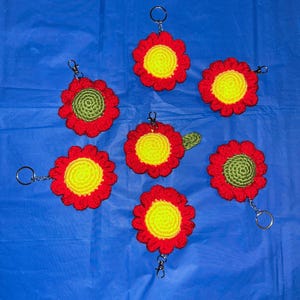 Puede incluir: Colección de siete llaveros de flores hechos a mano con ganchillo. Cada llavero presenta una flor roja con centro amarillo y un llavero plateado. Algunas flores tienen centros verdes. Los llaveros están sobre una superficie azul.