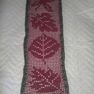 Puede incluir: Un camino de mesa rectangular hecho a mano con ganchillo. El camino de mesa presenta un diseño floral burdeos con un borde marrón oscuro. El patrón floral incluye hojas y flores estilizadas. El camino de mesa está colocado sobre una superficie blanca texturizada.