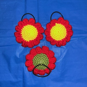 Puede incluir: Tres coleteros de flores de ganchillo hechos a mano. Cada flor presenta un diseño de pétalos rojos con un centro amarillo o verde. Los coleteros tienen una banda elástica negra. Los artículos se muestran sobre un fondo azul.