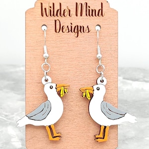 K&ouml;nnte beinhalten: Ein Paar Holzohrringe in Form einer M&ouml;we mit gelbem Schnabel und silbernen Haken. Die Ohrringe werden auf einer Holzkarten mit dem Text "Wilder Mind Designs" pr&auml;sentiert.