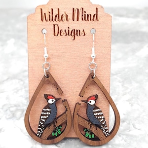Puede incluir: Pendientes de madera en forma de lágrima con diseño de pájaro carpintero. Los pendientes tienen un marco marrón con una ilustración detallada de un pájaro carpintero posado en una rama con hojas verdes. El texto "Wilder Mind Designs" es visible.
