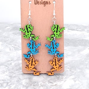 May include: A pair of dangle earrings featuring three stacked frog charms. The frogs are painted in shades of green, blue, and orange. The earrings are on a wooden display card with the text "Wilder Mind Designs".
