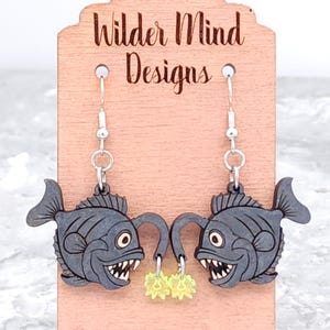 May include: A pair of dangle earrings featuring a black anglerfish with a glowing lure. The earrings are made of wood and have silver hooks. The earrings are displayed on a wooden card with the text "Wilder Mind Designs".