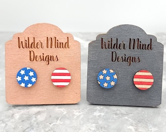 May include: Two pairs of wooden earrings with American flag designs. One pair features red and white stripes, the other features blue and white stars. The earrings are displayed on wooden stands with the text "Wilder Mind Designs" on each stand.