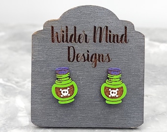 May include: A pair of green and purple poison bottle earrings with a skull and crossbones design. The earrings are on a gray wooden display card with the text "Wilder Mind Designs".