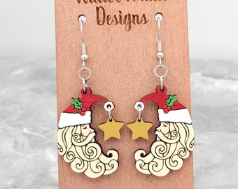 May include: A pair of wooden Christmas earrings featuring a Santa Claus face with a red hat and white beard, each with a dangling gold star. The earrings are on a wooden display stand with the text "Wilder Mind Designs".