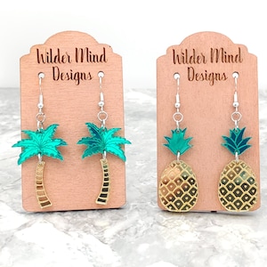 May include: Two pairs of dangle earrings on display cards. One pair features green palm trees with gold trunks. The other pair features gold pineapples with green tops.