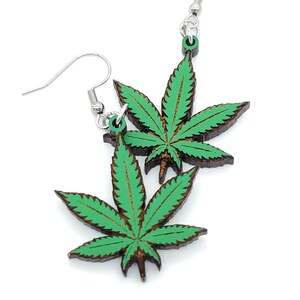 May include: A pair of green marijuana leaf earrings with silver hooks. The leaves are made of wood and have a dark brown outline.
