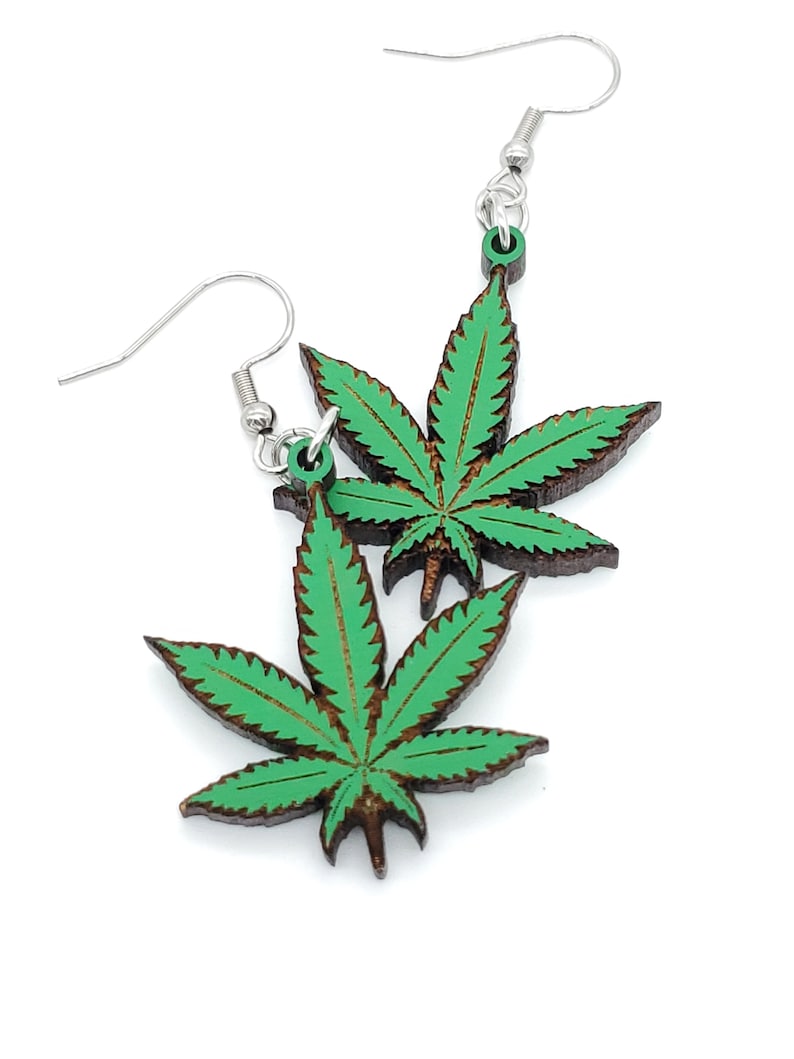 May include: A pair of green marijuana leaf earrings with silver hooks. The leaves are made of wood and have a dark brown outline.