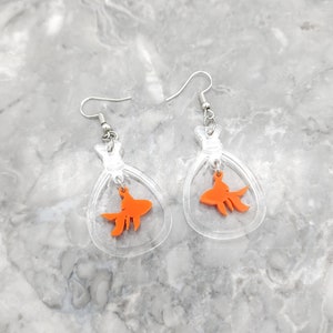 May include: A pair of clear acrylic earrings with a dangling orange goldfish charm. The earrings are shaped like a small bag or pouch.