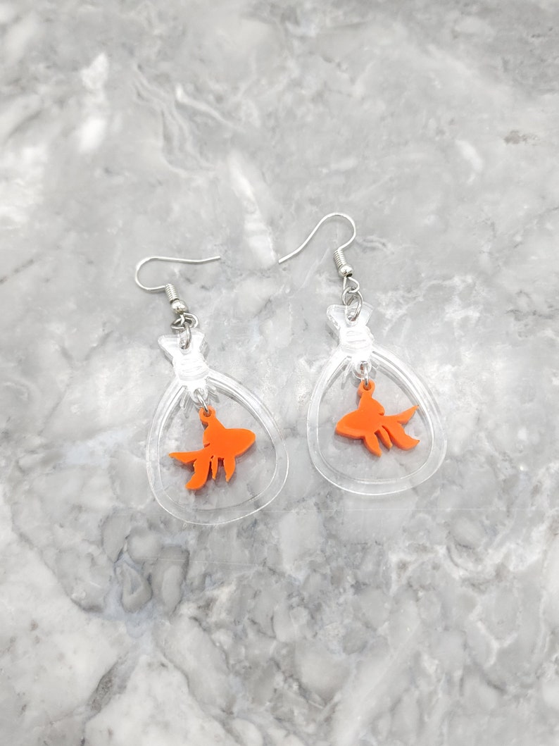 May include: A pair of clear acrylic earrings with a dangling orange goldfish charm. The earrings are shaped like a small bag or pouch.