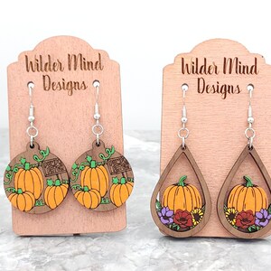 May include: Two pairs of wooden earrings with a fall theme. One pair features a round design with pumpkins and a "Pumpkin Patch" sign. The other pair features a teardrop design with a pumpkin and flowers.