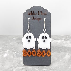 May include: A pair of white ghost earrings with orange glitter "BOO" charms hanging from them. The earrings are displayed on a black card with the text "Wilder Mind Designs".