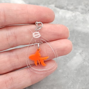 May include: A clear acrylic charm with a small orange goldfish inside. The charm is attached to a silver jump ring.