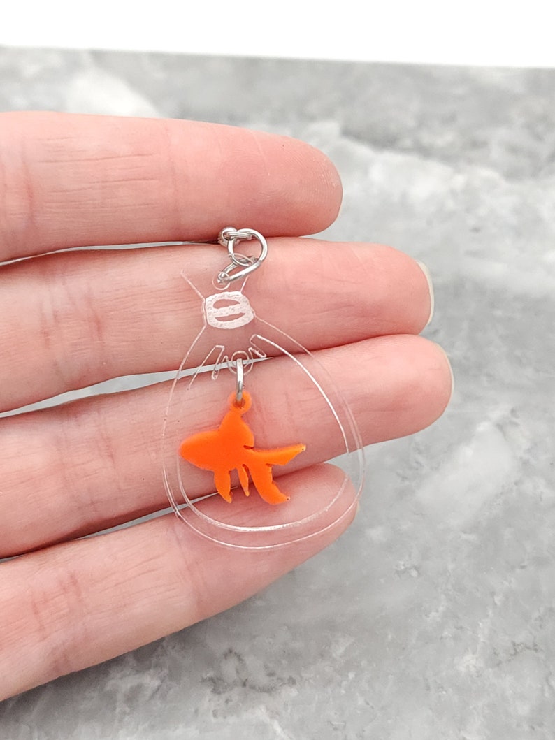 May include: A clear acrylic charm with a small orange goldfish inside. The charm is attached to a silver jump ring.