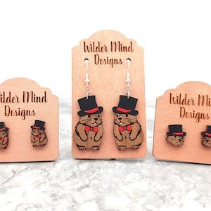 May include: Three pairs of wooden earrings featuring a cartoon groundhog wearing a top hat and bow tie. The earrings are displayed on wooden stands with the text "Wilder Mind Designs" engraved on each stand.