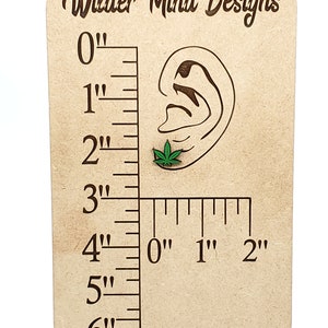 May include: A wooden ruler with a carved ear and a green marijuana leaf earring. The ruler measures up to 6 inches.