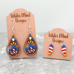 May include: Two pairs of wooden earrings with a red, white, and blue firework design. The larger pair is a teardrop shape with a silver hook. The smaller pair is a stud with a silver post. The earrings are displayed on a wooden stand with the text "Wilder Mind Designs".