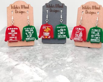 May include: A set of six pairs of Christmas sweater earrings. Each pair features a different design with a humorous holiday theme. The earrings are made of red, green, and white acrylic and have silver hooks.