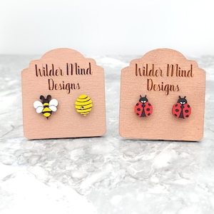 May include: Two pairs of wooden earrings on display cards. The first pair features a bee and a honeycomb. The second pair features two ladybugs. The cards are labeled "Wilder Mind Designs".