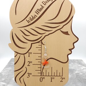 May include: Clear glass dangle earrings with a small orange goldfish inside. The earrings are hanging from a wooden mannequin head with a ruler behind it. The ruler shows the earrings are 1 inch long.