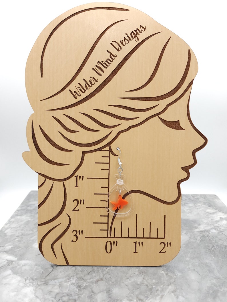 May include: Clear glass dangle earrings with a small orange goldfish inside. The earrings are hanging from a wooden mannequin head with a ruler behind it. The ruler shows the earrings are 1 inch long.