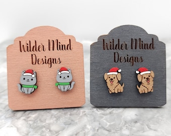 Puede incluir: Dos pares de pendientes de madera con un gato y un perro con gorros de Papá Noel. El gato es gris con una bufanda verde y el perro es marrón. Los pendientes se muestran en dos tarjetas de madera con el texto "Wilder Mind Designs".