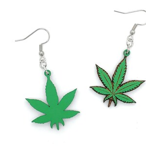 May include: A pair of green marijuana leaf earrings with silver hooks. The leaves are made of wood and have a detailed design.