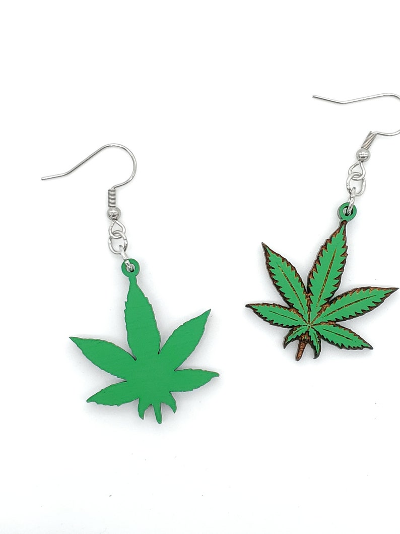 May include: A pair of green marijuana leaf earrings with silver hooks. The leaves are made of wood and have a detailed design.
