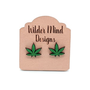 May include: A pair of wooden earrings featuring a green marijuana leaf design. The earrings are displayed on a wooden card with the text "Wilder Mind Designs".