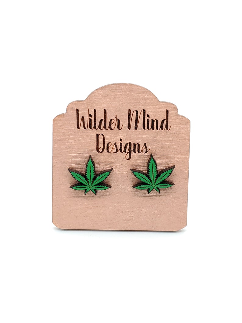 May include: A pair of wooden earrings featuring a green marijuana leaf design. The earrings are displayed on a wooden card with the text "Wilder Mind Designs".