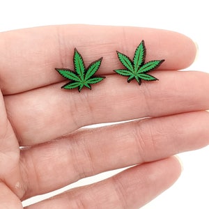 May include: A pair of green marijuana leaf earrings. The earrings are made of wood and have a dark brown color.