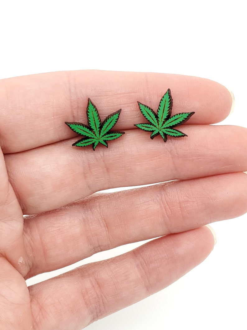 May include: A pair of green marijuana leaf earrings. The earrings are made of wood and have a dark brown color.