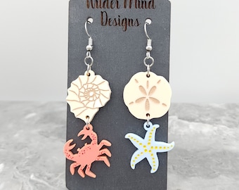 May include: A pair of dangle earrings featuring a seashell, starfish, and crab design. The earrings are made of wood and painted in shades of blue, yellow, pink, and white. The earrings are hanging from silver hooks. The card behind the earrings reads "Wilder Mind Designs".