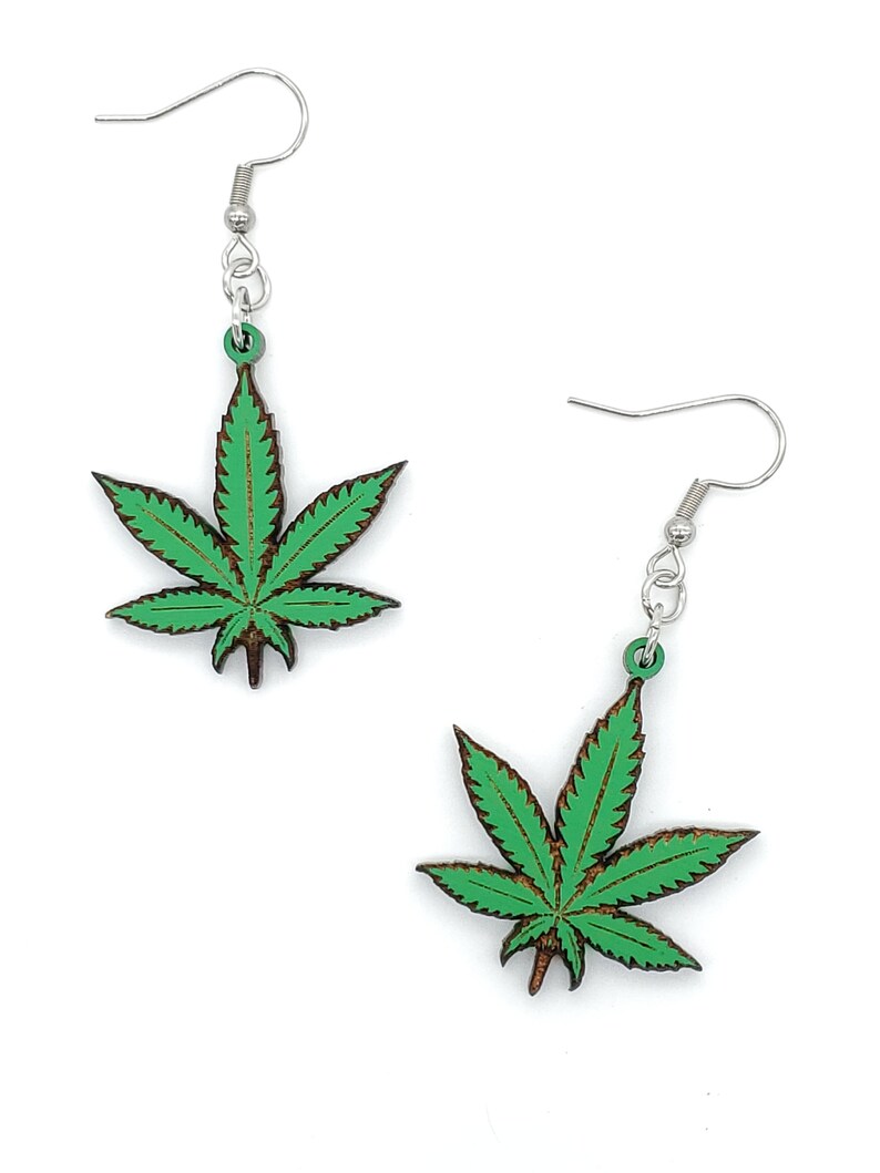 May include: A pair of green marijuana leaf earrings with silver hooks. The leaves are made of wood and have a dark brown outline.