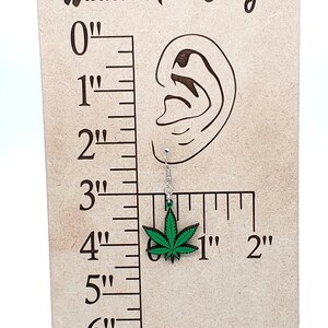 May include: Green marijuana leaf earrings on a wooden display with a ruler. The earrings are 1 inch long.