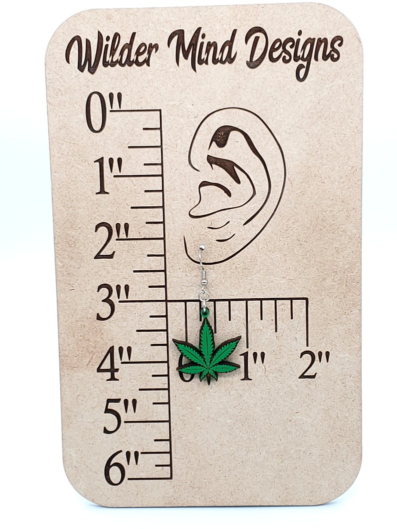 May include: Green marijuana leaf earrings on a wooden display with a ruler. The earrings are 1 inch long.